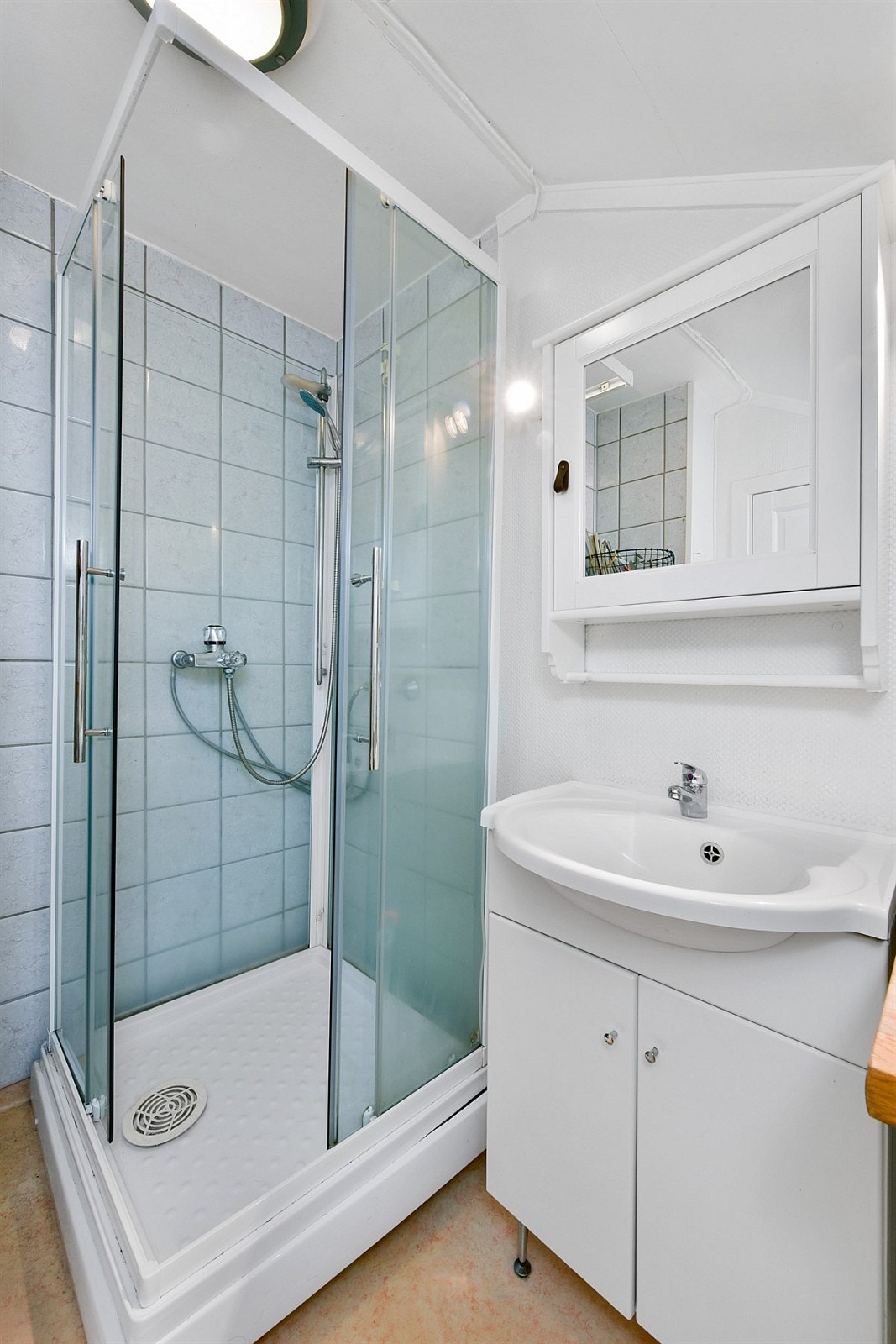 På badet er plassen maksimalt utnyttet. Rommet er innredet med sandfarget gulvbelegg, hvite, tapetserte vegger, samt blå veggfliser i dusjsonen. Galleribilde