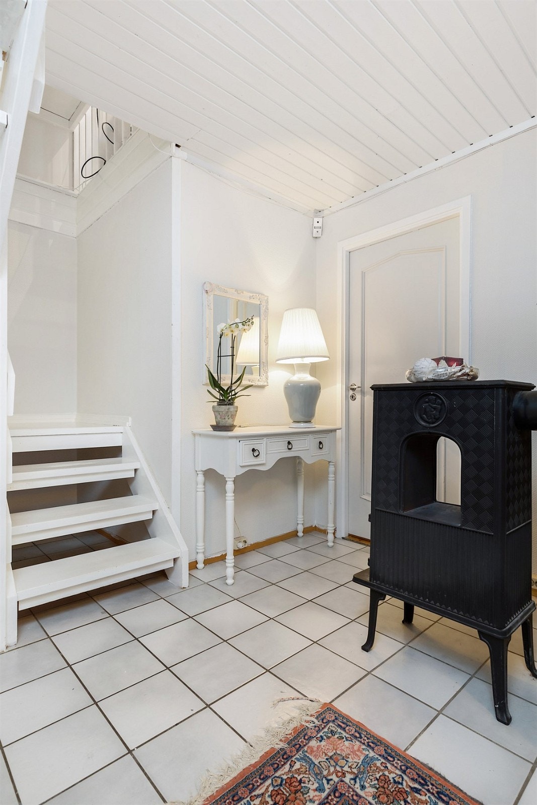 Gangen har flislagt gulv, lysmalte vegger og en åpen trapp som leder opp til første etasje. Rommet har plass til entrémøblement og en koselig vedovn sørger for ekstra varme på kalde dager. Galleribilde