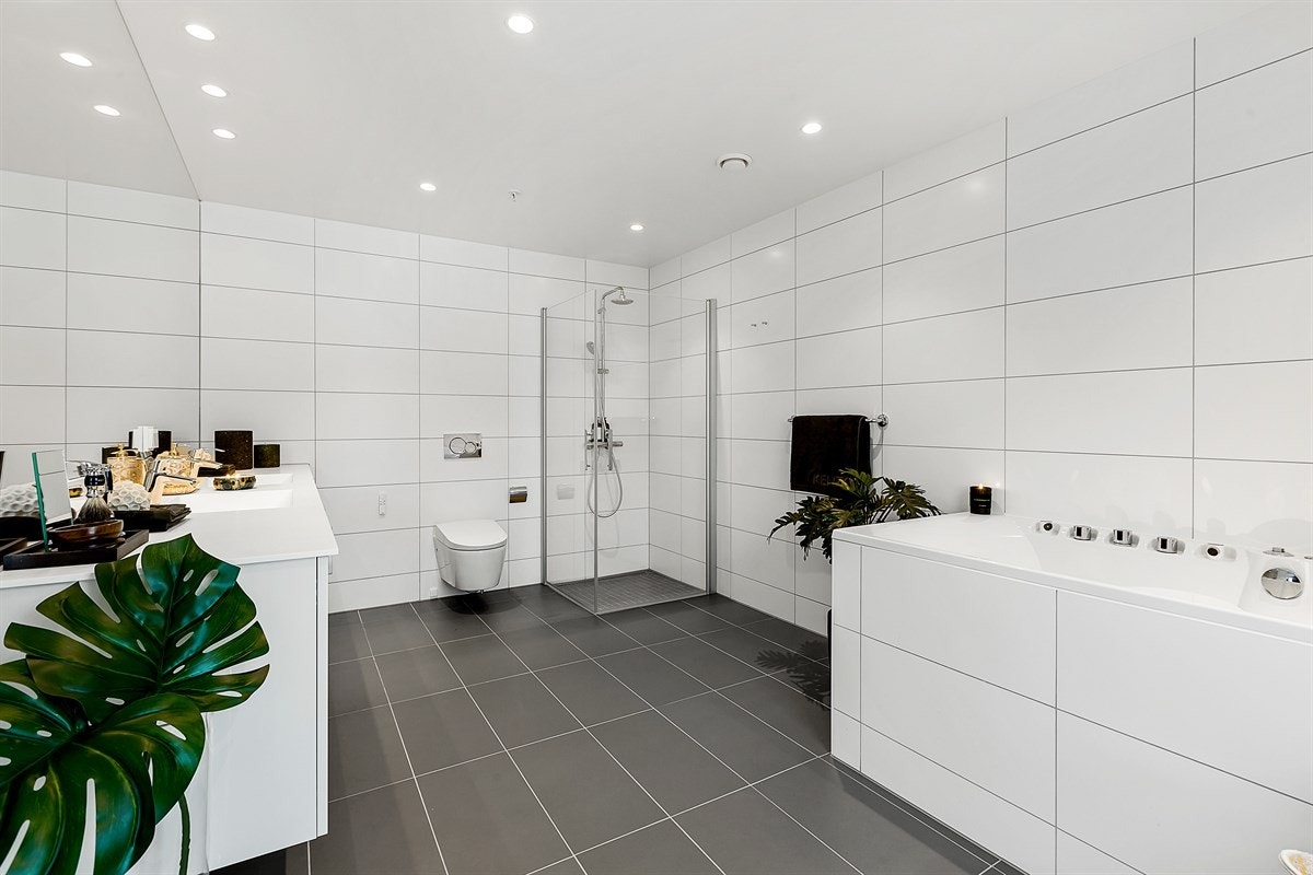 Badet har gulvfliser med gulvvarme, hvite veggfliser og downlights i tak. Videre er badet utstyrt med vegghengt toalett, stort speil og bred skapseksjon med heldekkende dobbelservant. Galleribilde