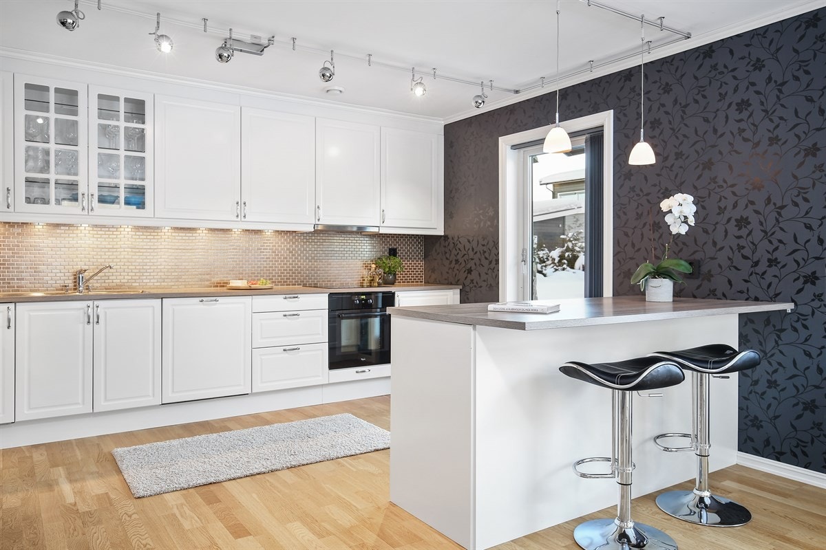 Kjøkkenet ligger åpent i stuen med barløsning, noe som skaper en sosial og god romfølelse. Rommet har parkett på gulv, vegger med dekorativt tapet og dør til en overbygd markterrasse Galleribilde