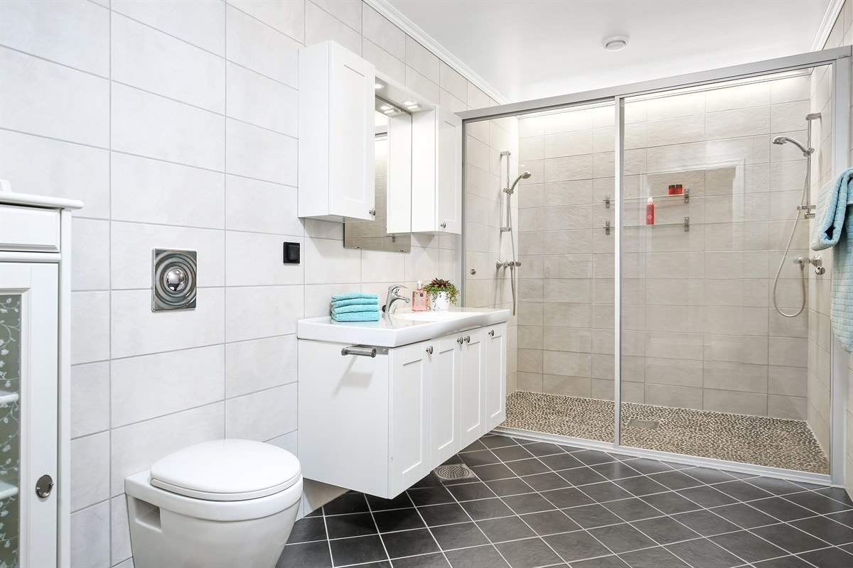 Det ene badet har lyse veggfliser og sorte gulvfliser, i kombinasjon med sjøstein i en dusjnisje med dobbel dusj. Galleribilde