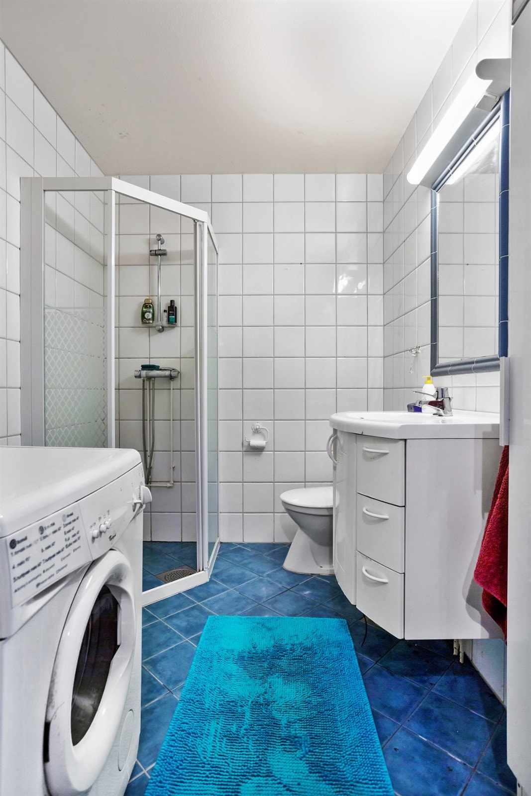 Bad med veggmonterte dusjvegger, klosett, servant med skapinnredning og opplegg for vaskemaskin. Galleribilde