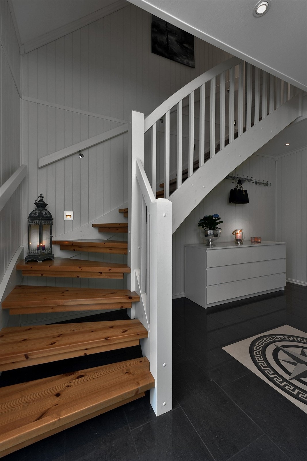 Under en trapp med hvite vanger og eiketrinn er det åpen garderobeløsning. Hallen er praktisk tilknyttet et bad og et teknisk rom/bod, og en glassdør fører inn til en romslig stue. Galleribilde