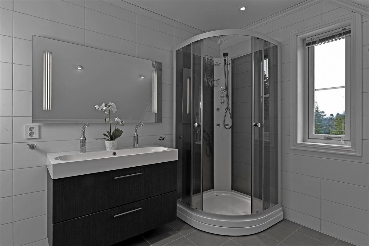 Rommet er innredet med heldekkende servant på romslig skuffeseksjon i mørk utførelse, høyskap med speilfront og speil med belysning over servant. Galleribilde