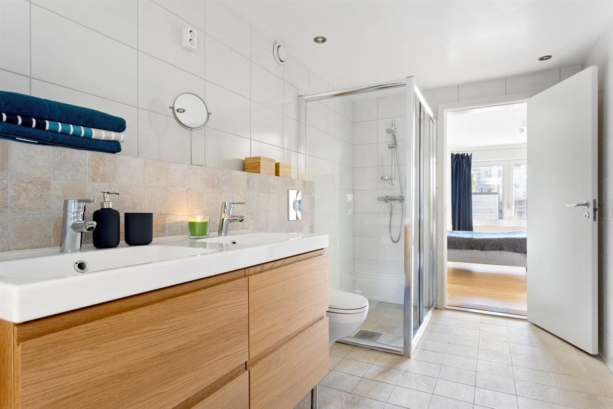 Romslig bad med fliser på gulv og vegger, samt gulvvarme. Badet består av åpen dusjplass med dusjhjørne, dobbel heldekkende servant i skapinnredning, vegghengt wc og opplegg for vaskemaskin. Galleribilde