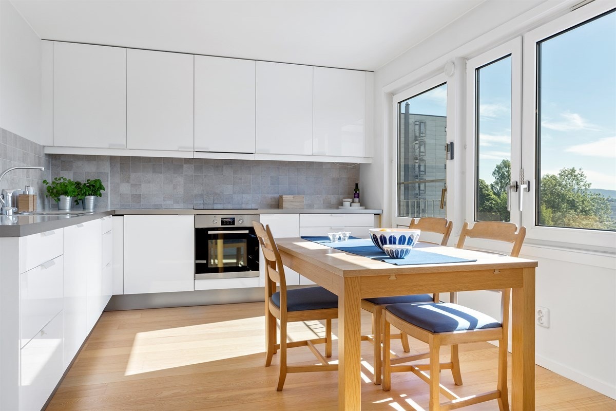 Kjøkkeninnredning med hvite fronter, enkel oppvaskkum, fliser mellom overskap og laminert benkeplate. Kjøkkenet er utstyrt med ettgrepskran, integrert oppvaskmaskin, kjøleskap, frys, og komfyr med koketopp. Galleribilde