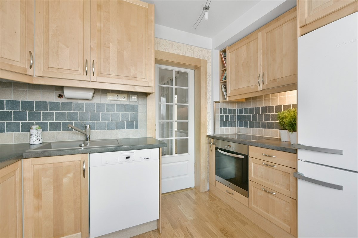 Innredningen har tradisjonelle profilfronter i naturfarge, vitrineskap og belysning over laminert benkeplate med vaskekum. Galleribilde