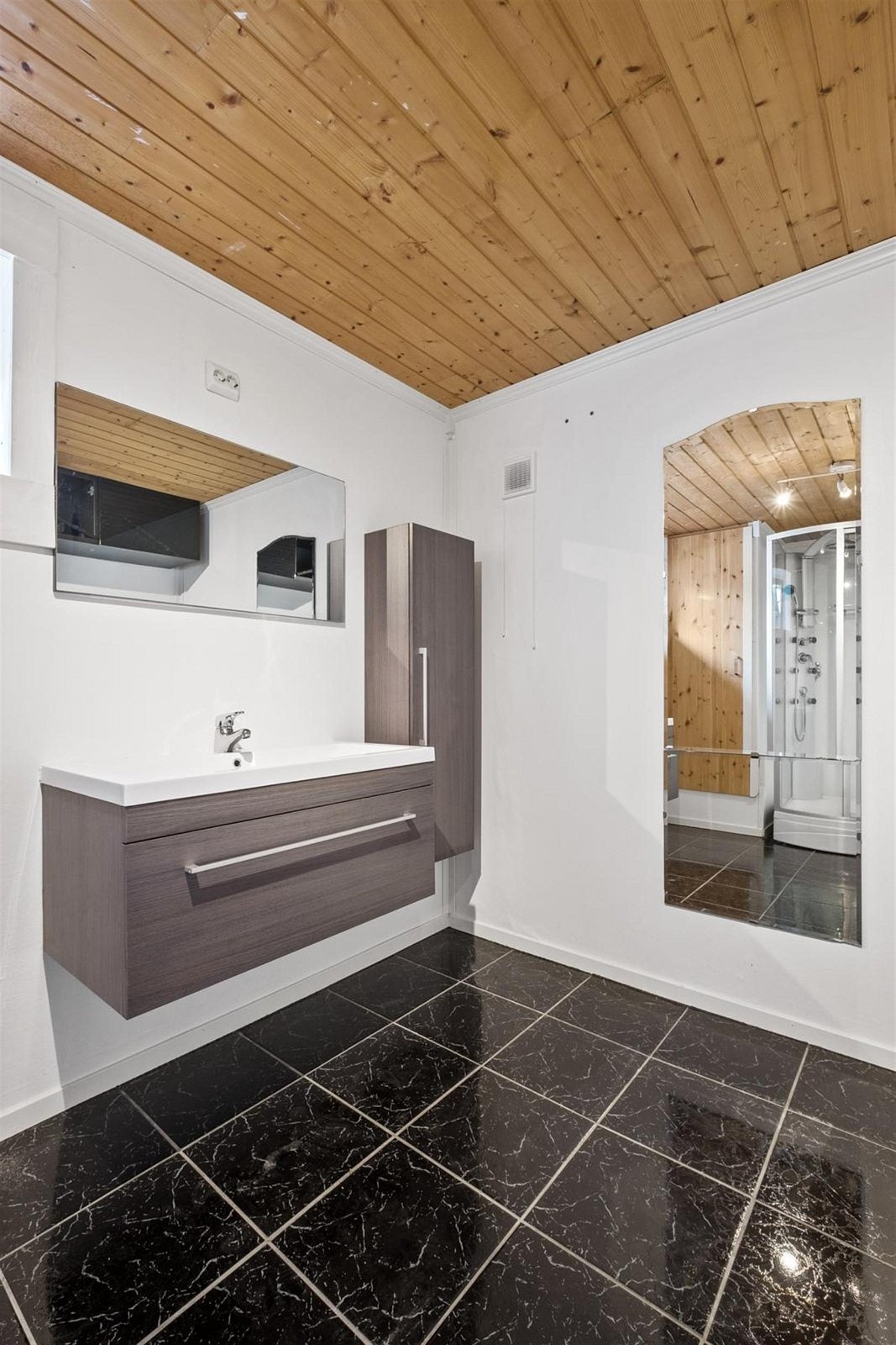 Bad i underetasje har mørkefliser på gulv og lysmalte vegger. Innredningen er stilig med skuff under servant og matchende høyskap. Badet har også dusjkabinett. Galleribilde