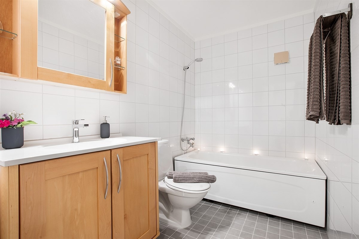 Delikat og tidløst flislagt bad med servant med underskap, overskap med speilfront, frittstående klosett, badekar og vegghengt håndkletørker. Galleribilde