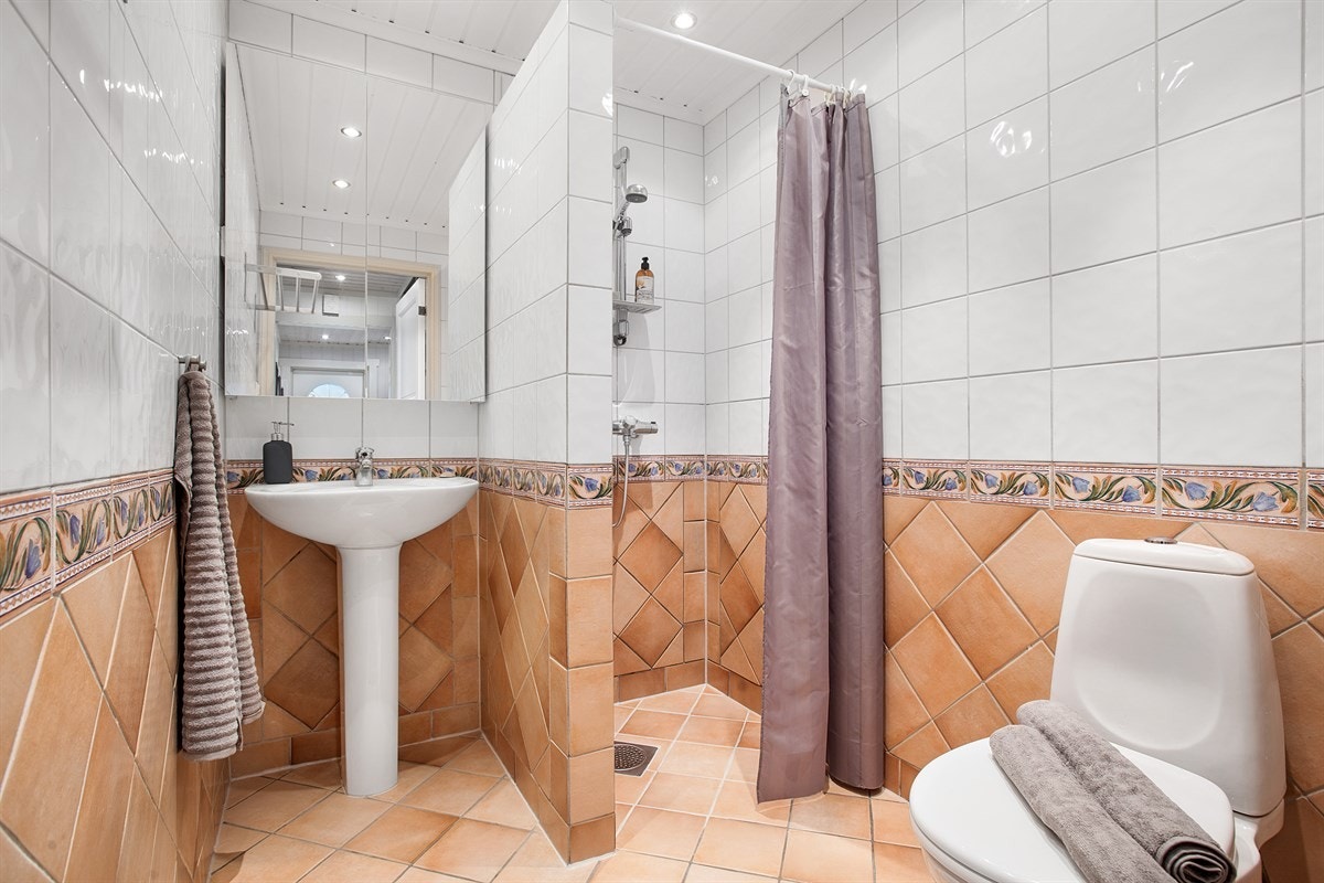 Delikat flislagt bad med varmekabler i gulv, frittstående toalett, servant, overskap med speilfront, dusjnisje og vegghengt håndkletørker. Galleribilde