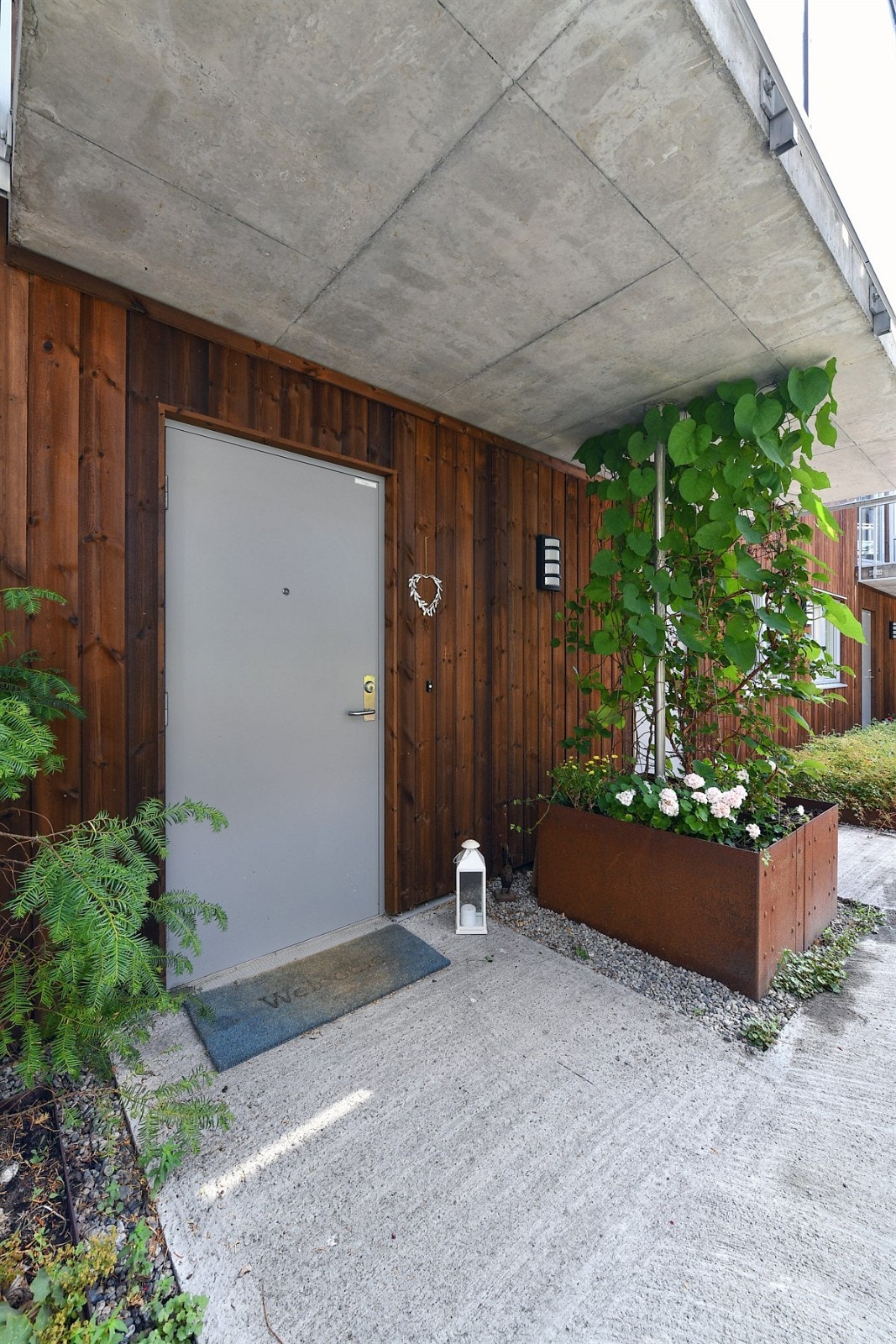 Fra felles atrium, under verandaen i andre etasje, har boligen et pent inngangsparti med utelys, støpt dekke og frodige blomsterkasser som bidrar til grønn skjerming. Galleribilde