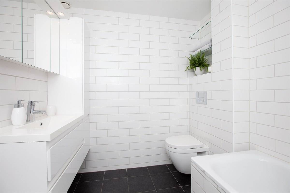 Badet utstyrt med vegghengt toalett, hvit høyglanset innredning med heldekkende servant og innmurt flislagt badekar med dusj og sprutdemper Galleribilde