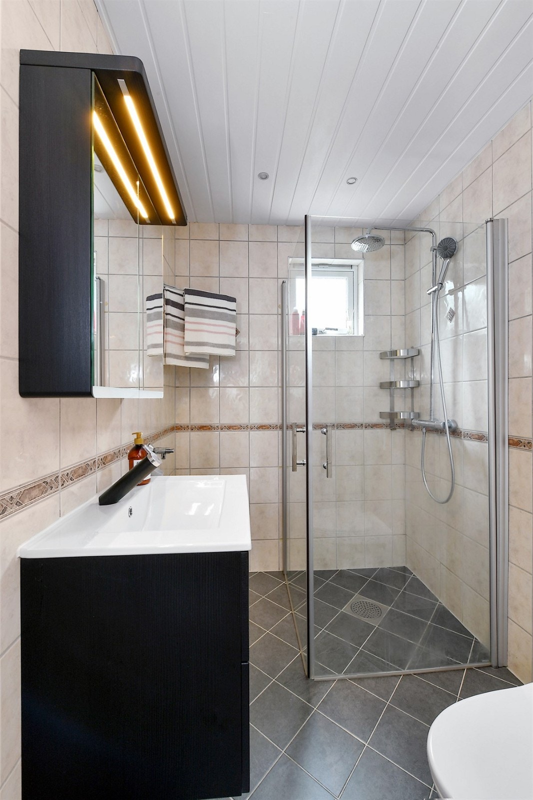 Badet er pent innredet med gulv- og veggfliser, gulvvarme, downlights og vegghengt toalett. Galleribilde