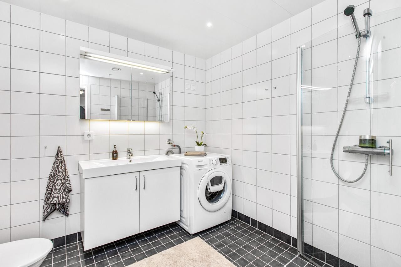 Flislagt bad med sammenleggbare dusjdører, vegghengt toalett, romslig innredning og opplegg for vaskemaskin. Galleribilde