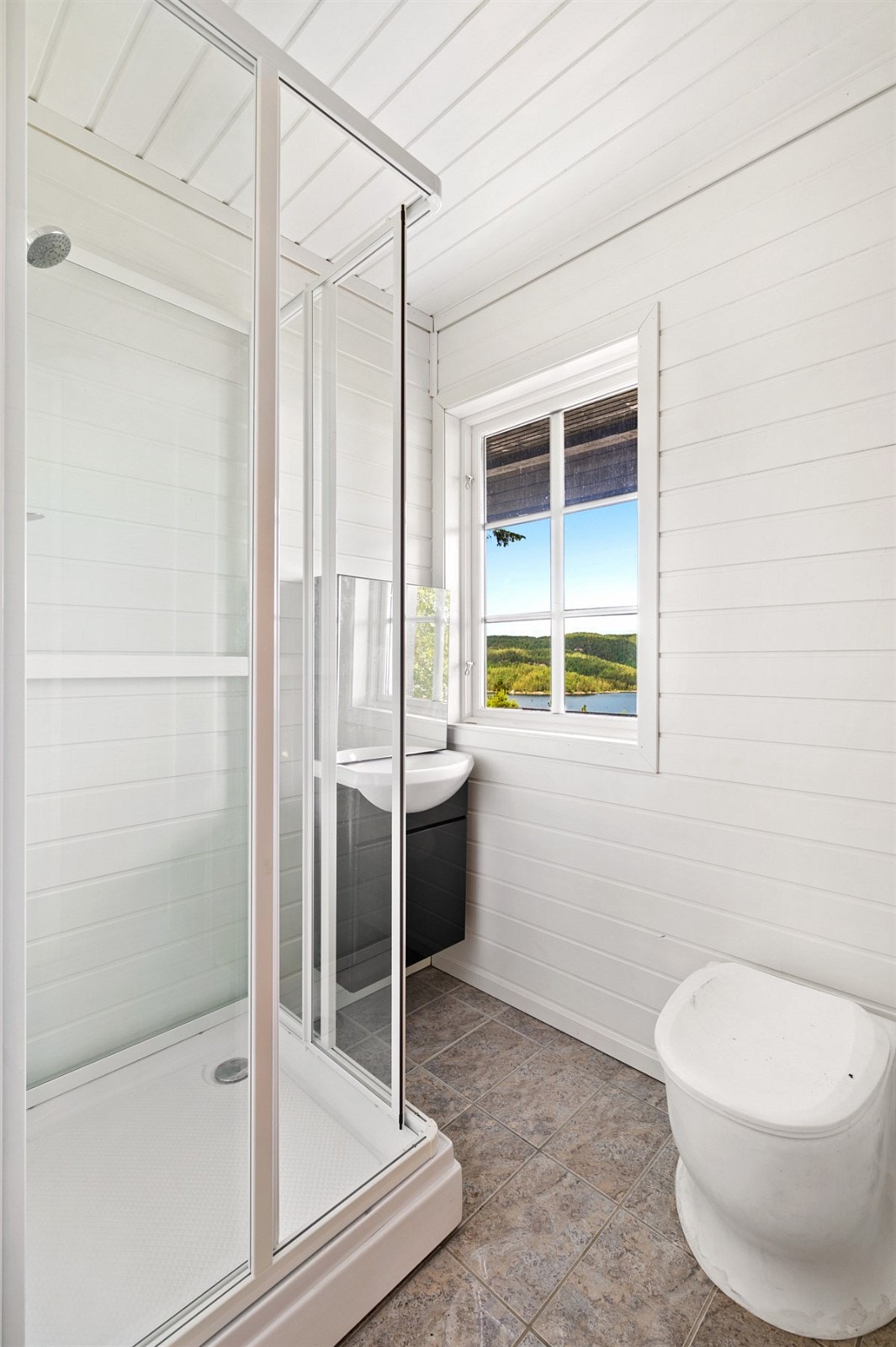 Badet er utstyrt med servant, dusjkabinett og toalett. Det er flis på gulv og lysmalt panel på veggene. Galleribilde