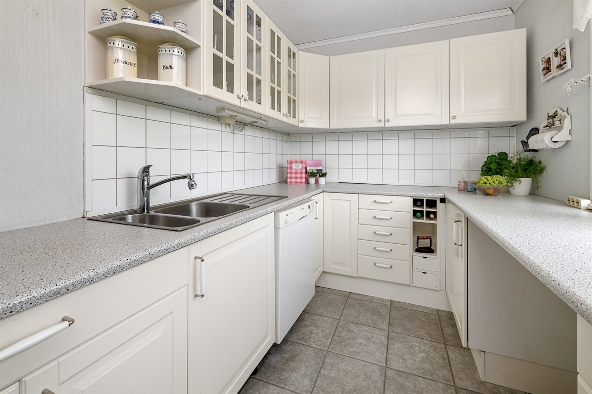 Benkeplaten er i grå laminat med en nedfelt dobbel oppvaskkum, mens veggen ovenfor er kledd med plater i fliseimitasjon. Videre har kjøkkenet benkebelysning og flere frittstående hvitevarer. Galleribilde