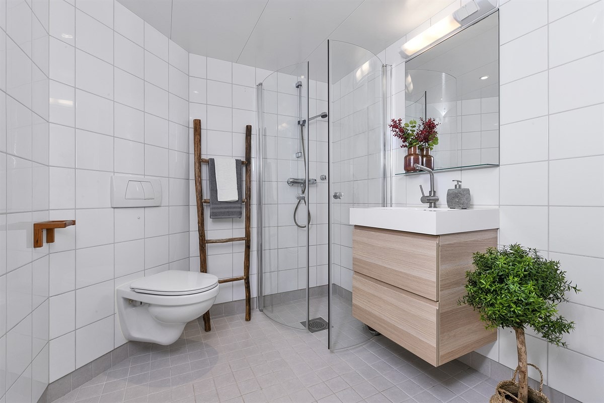 Badet fremstår som pent med hvite veggfliser, grå gulvfliser og behagelig gulvvarme. Innredningen fra 2015 har naturfargede skuffefronter og heldekkende servant, samt speil med glasshylle og belysning over. Galleribilde