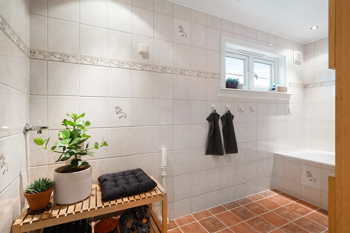 Videre har badet innfliset badekar med dusj og dusjdør i glass. Det er baderomsinnredning med servant og opplegg for vaskemaskin. Galleribilde