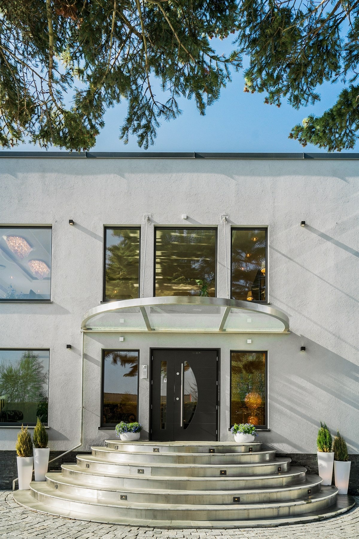 PROAKTIV Eiendomsmegling - Inngangspartiet har en halvmåneform med flislagte trinn med spesialkonstruerte stålbeslag, varme og belysning, samt et takoverbygg i samme fasong. De avrundede konstruksjonene bryter stilsikkert opp den stramme og rette fasaden. Galleribilde