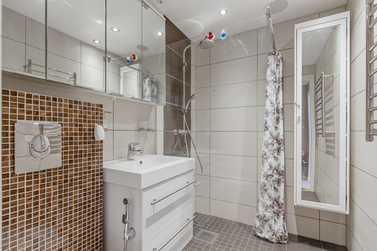 Badet består av vegghengt wc, servant med underskap, overskap med speilfronter og overlys, veggskap, samt åpen dusj med glassvegg og forheng. Galleribilde