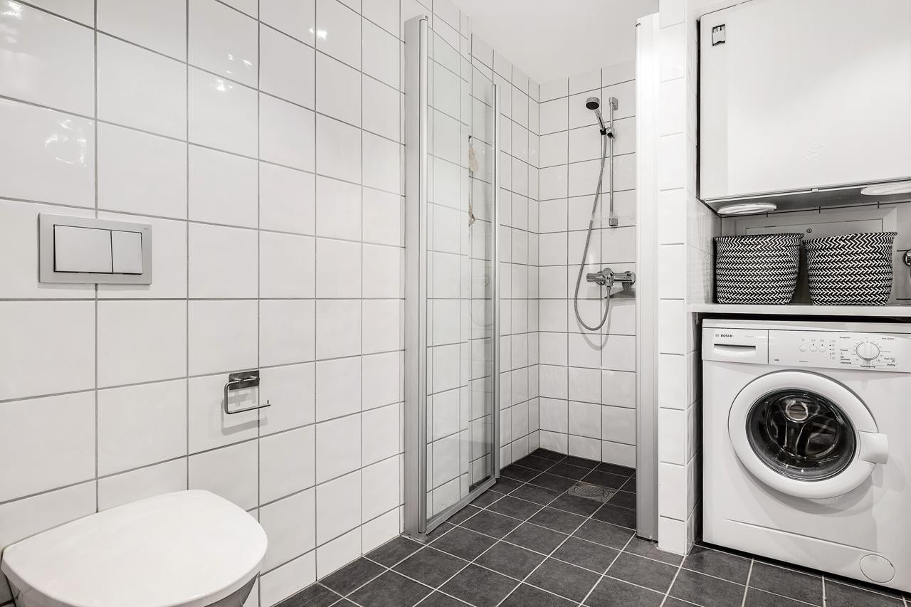 Stort flislagt bad med dusjhjørne, veggfestet wc, hvite baderomsmøbler og opplegg for vaskemaskin og tørketrommel Galleribilde