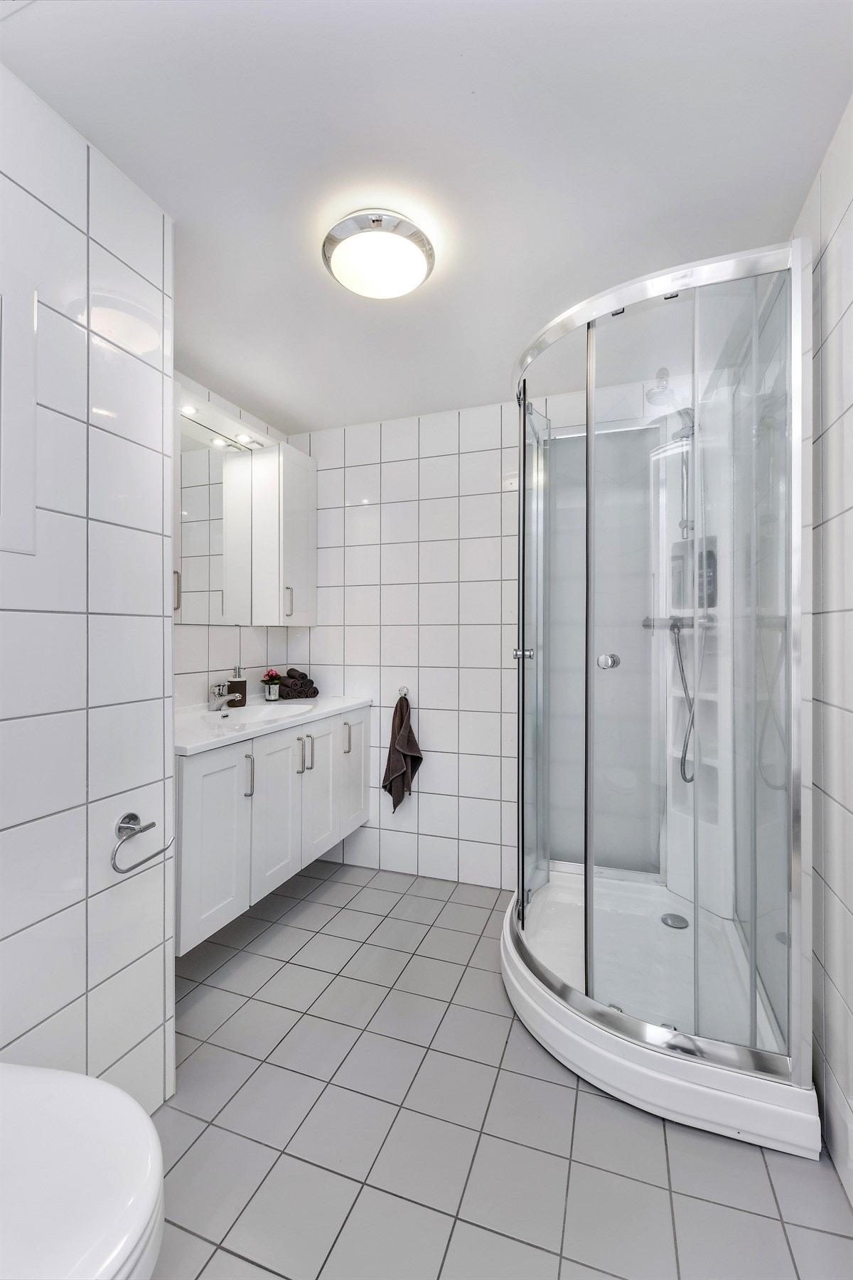 Badet gir et tidløst inntrykk, med hvite veggfliser og grå gulvfliser med varme. Innredningen består av en heldekkende servant på underskap med hvite, profilerte fronter, samt matchende veggskap og et speil med belysning. Galleribilde