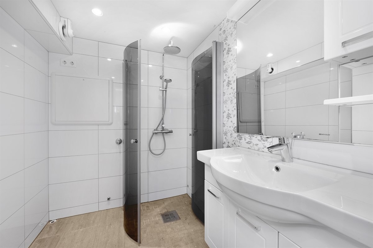 Badet i underetasjen har flislagte overflater, gulvvarme, downlights og fuktstyrt vifte. Innredningen har hvite, profilerte fronter, heldekkende servant, veggskap med hylle i underkant, speil og belysning. Galleribilde