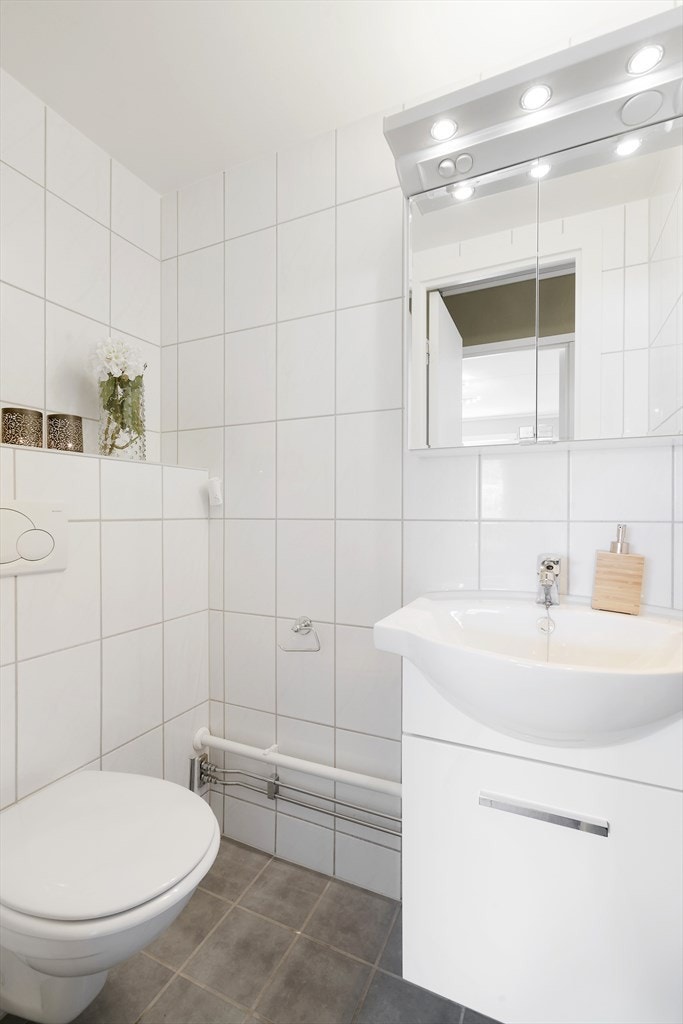 Flislagt toalettrom fra 2010 i lyse farger, servantskap med dører i matt hvit utførelse, og ovenpåliggende servant av porselen. Speilskap med belysning og stikkontakt på vegg over servant, samt. vegghengt toalett. Galleribilde