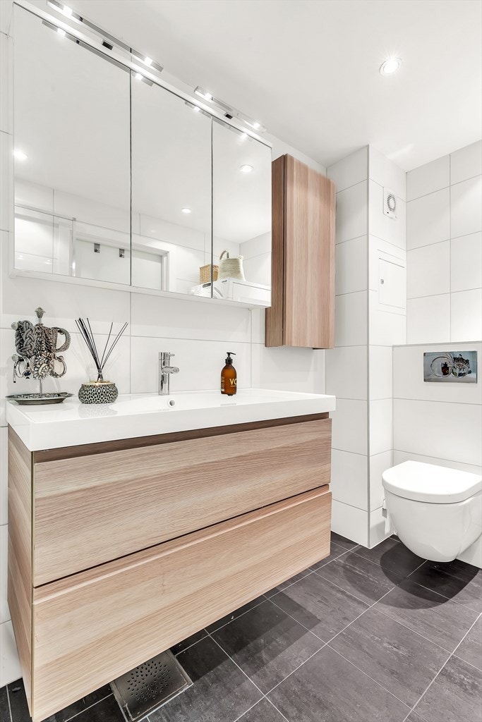 Badet er flislagt og har god belysning fra downlights med dimmer.
Hel servantplate med servantskap. Overskap med speildører. Veggfestet toalett med skjult sisterne og softclose sete. Innmurt badekar med glass dusjvegg. Galleribilde