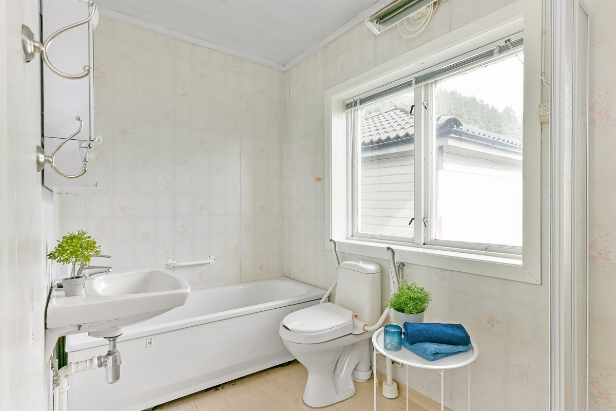 Badet har vinyl på gulv og vegger, og trenger oppussing for å møte dagens krav til standard. Rommet er funksjonelt, og innredet med servant, enkel innredning, gulvmontert toalett, dusjkabinett og badekar. Galleribilde