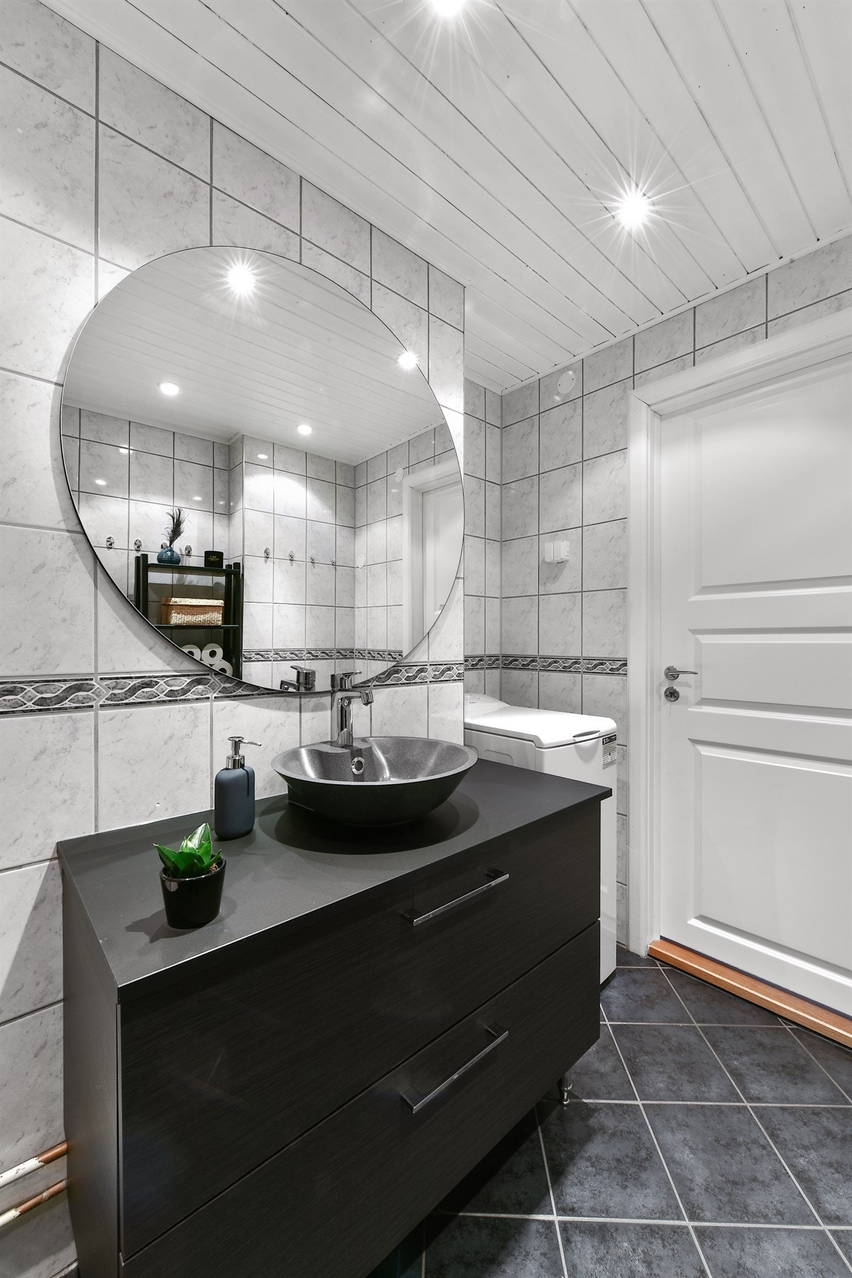 Videre er det et stort, rundt speil, et toalett og et dusjhjørne med buede glassdører. Badet er utstyrt med opplegg for vaskemaskin. Galleribilde