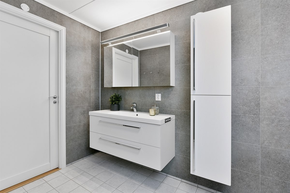 Badet er innredet med servant på skuffeseksjon med hvite fronter, matchende høyskap og speilskap med lys. Galleribilde