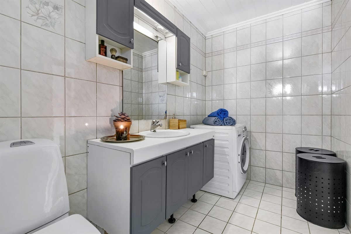 Det er innredet med servant på hvitt skap med blå, profilerte fronter, matchende overskap og speil med belysning. Videre har badet dusjkabinett, toalett og opplegg for vaskemaskin. Galleribilde