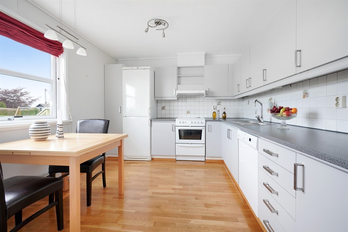 Kjøkken med originale fronter malt i lysegrå og laminat benkeplate. Utsikten fra kjøkkenet er mot sjøen og friområdet. Galleribilde