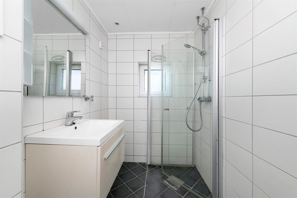 Rommet har gulvvarme, downlights og lysinnslipp fra vindu. Innredningen består av servantskap og speil med overlys. Videre er det vegghengt toalett, dusjhjørne med innfellbare glassdører og opplegg for vaskemaskin. Galleribilde