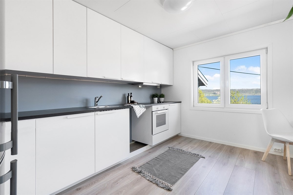 Innredningen har takhøye overskap med slette, hvite fronter. Benkeplaten er i mørk laminat med en nedfelt oppvaskkum. Veggen ovenfor er holdt i en blågrå nyanse. Galleribilde