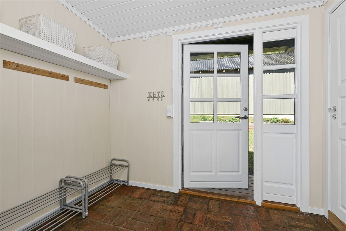 Entreen har rød teglstein på gulvet og lysmalte panelvegger. I tillegg er det to knaggrekker, en plassbygget hylle og dør inn til et praktisk gjestetoalett. Galleribilde