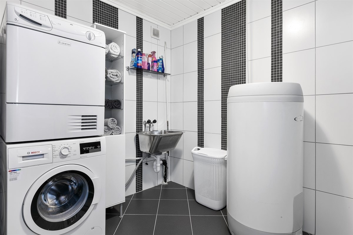 Vaskerommet ligger i forbindelse med en gang og har mørke gulvfliser og hvite veggfliser i kombinasjon med mørke mosaikkfliser. Galleribilde