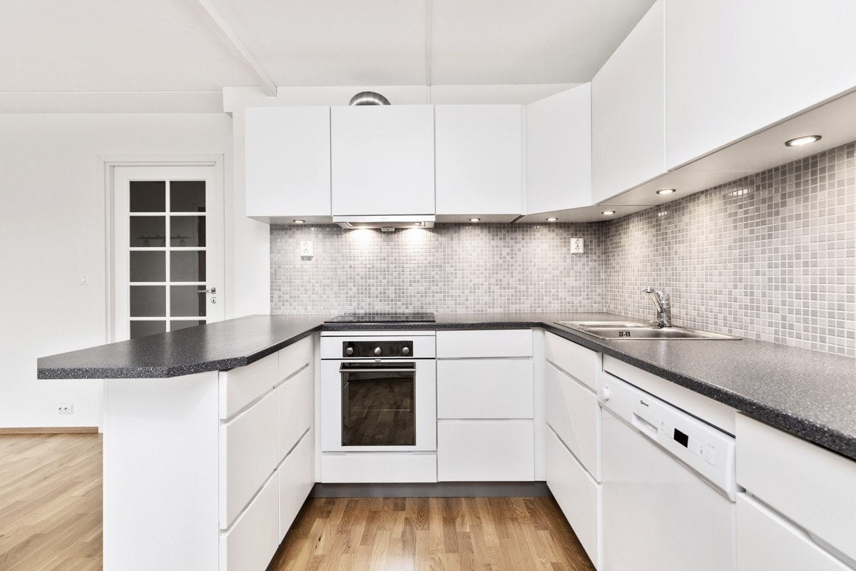 Tidsmessig kjøkkeninnredning med hvite glatte fronter, benkeplater i laminat, rustfri oppvaskkum, ventilator, opplegg for oppvaskmaskin og integrerte hvitevarer. Galleribilde