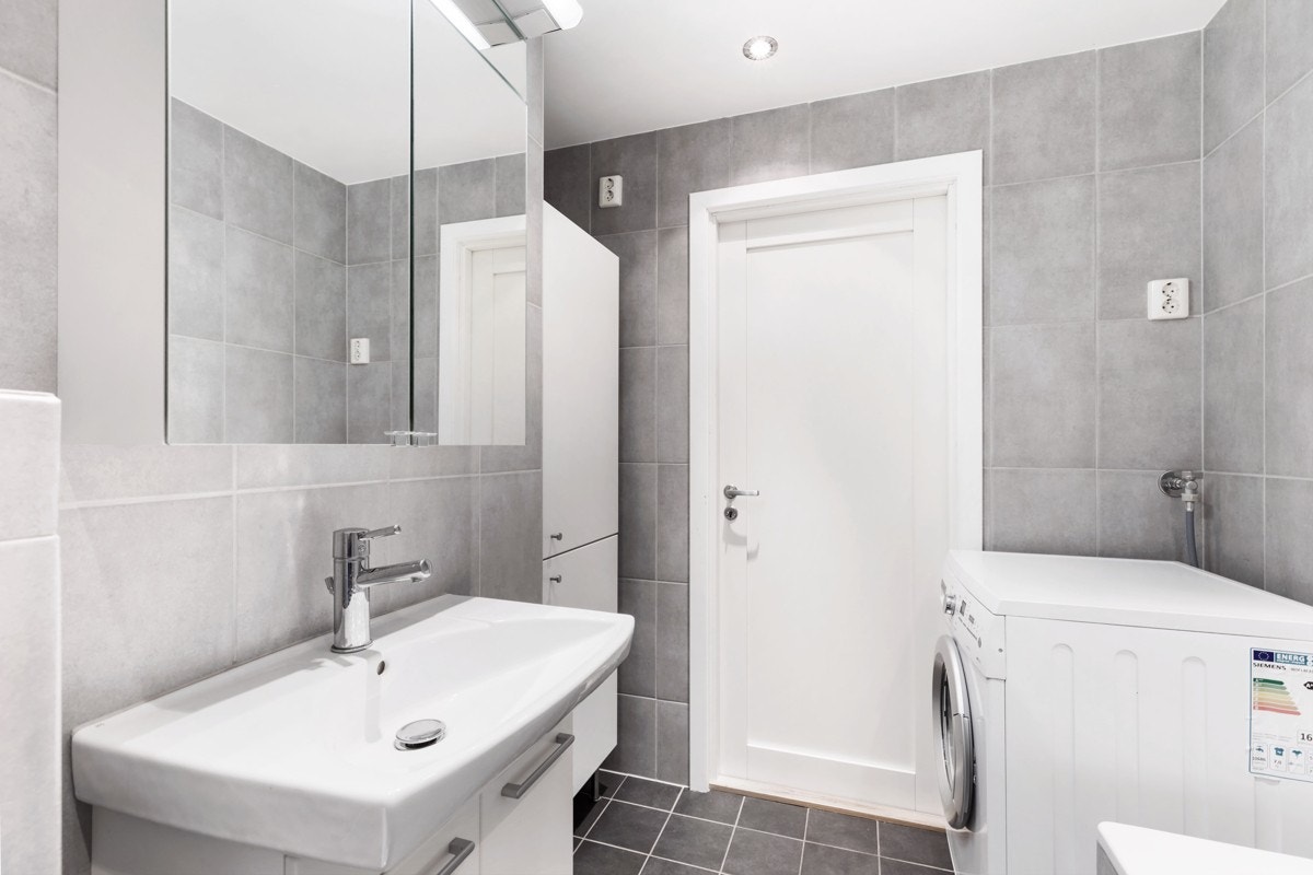 Badet inneholder veggmontert dusjvegg, innmurt badekar, klosett, servant med skuffer, speilskap og opplegg for vaskemaskin. Galleribilde