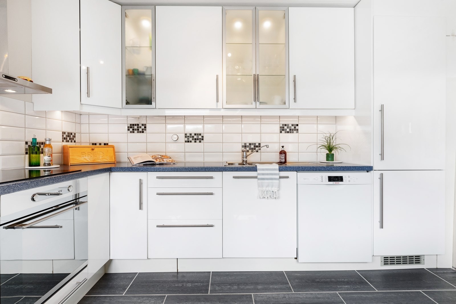 Nesten takhøy kjøkkeninnredning med høyglansede folierte fronter, 4 overskap med frostet glass og integrert lys, laminat benkeplate, fliser over benken, ventilator med stålhette, opplegg for oppvaskmaskin og stål benkebeslag. Galleribilde