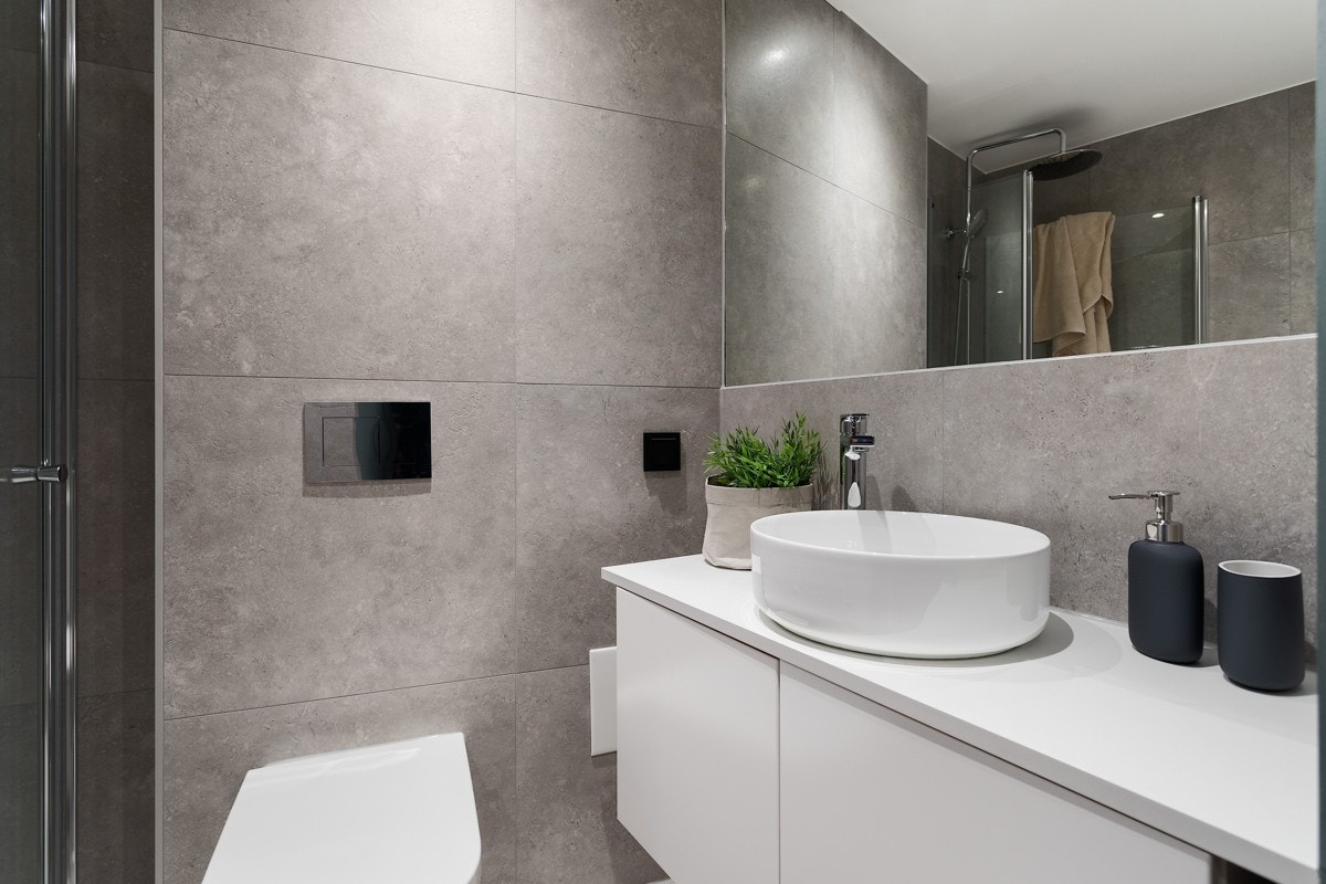 Lekkert bad med varmekabler, betongflis på gulv, mosaikk i dusjsonen, limt speil på hel vegg, dusj med svingbare dusjdører i glass, veggmontert wc, servant på benkeplate samt opplegg for vaskemaskin. Galleribilde