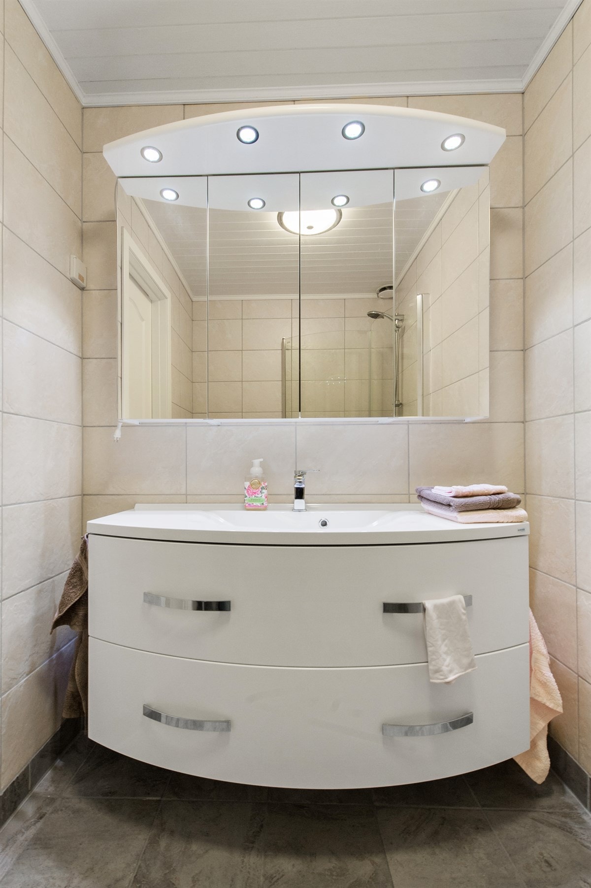 Fin og praktisk baderomsinnredningen bestående av stor servant med skuffer og dusjhjørne med buede glassdører Galleribilde