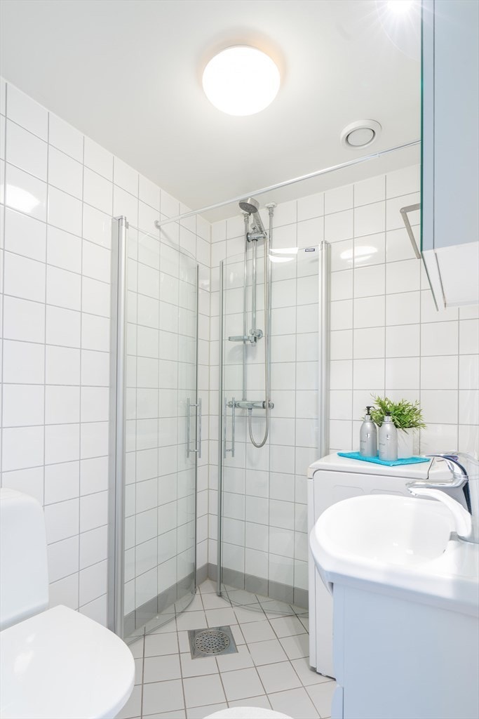 Flislagt baderom som består av gulvmontert toalett, dusjhjørne, opplegg for vaskemaskin, servantinnredning med skap under og speilskap med lys Galleribilde