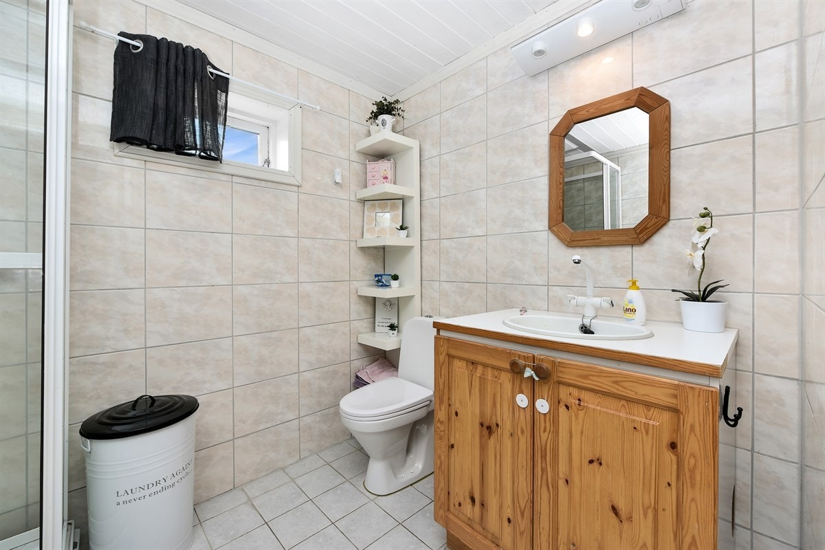 Badet er eldre, har flislagte overflater og er utstyrt med dusjkabinett, gulvmontert toalett og benkeskap med servant. Galleribilde