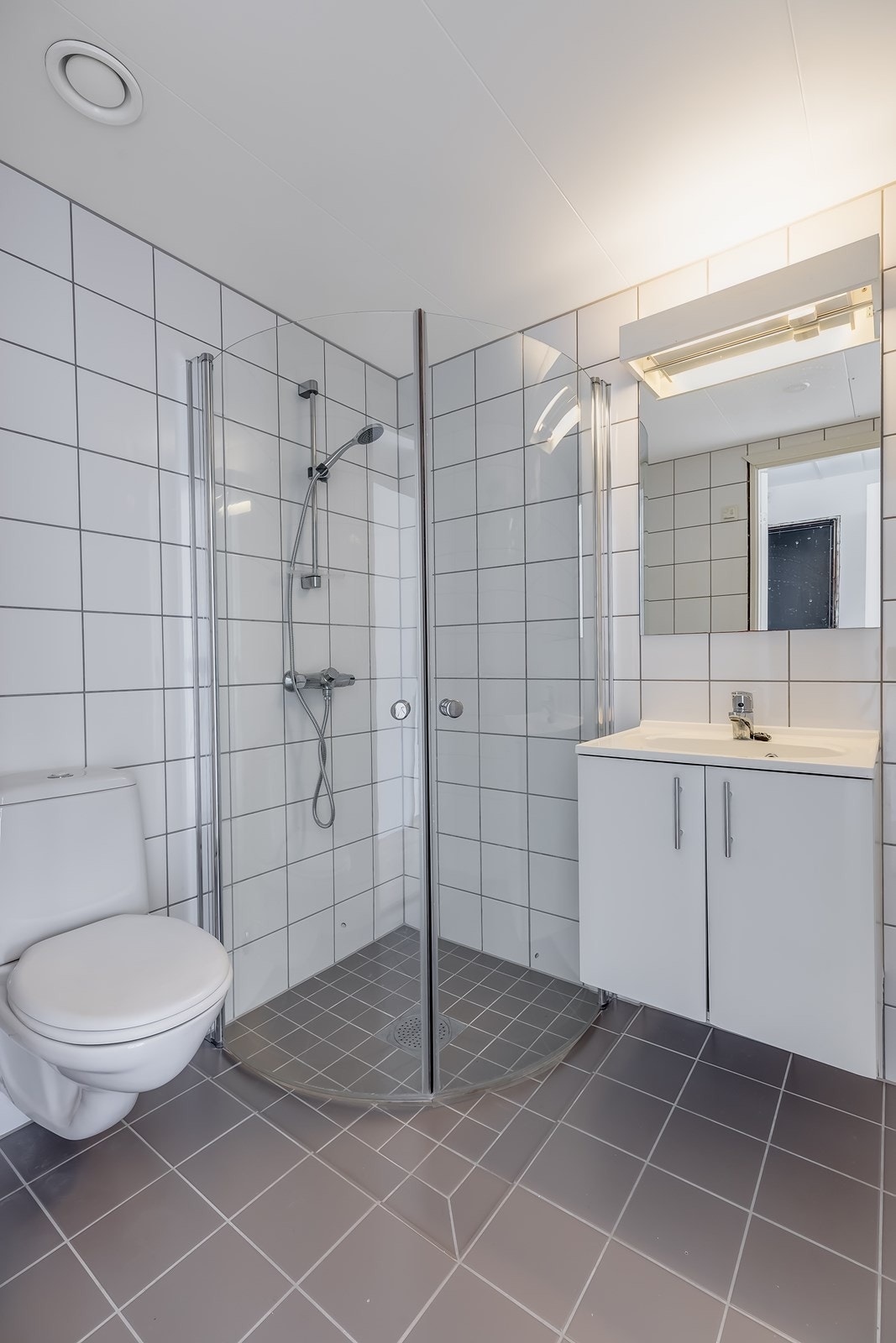 Badet inneholder vegghengt toalett, dusj med innfellbare dører og servant med underskap og speil over. Galleribilde