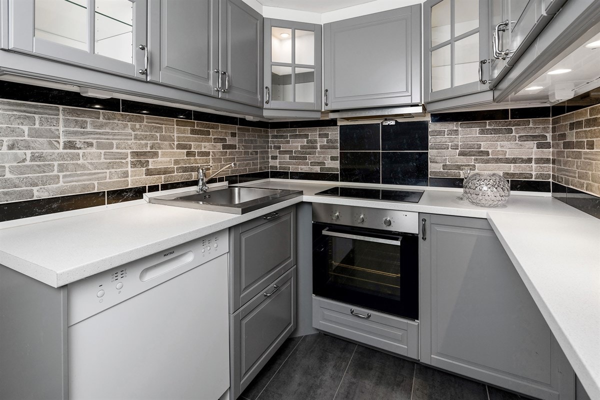 Kjøkkenet har grå, profilerte fronter, vitrineskap med belysning, benkeplate i lys laminat med nedfelt vaskekum og steinfliser over benk. Galleribilde