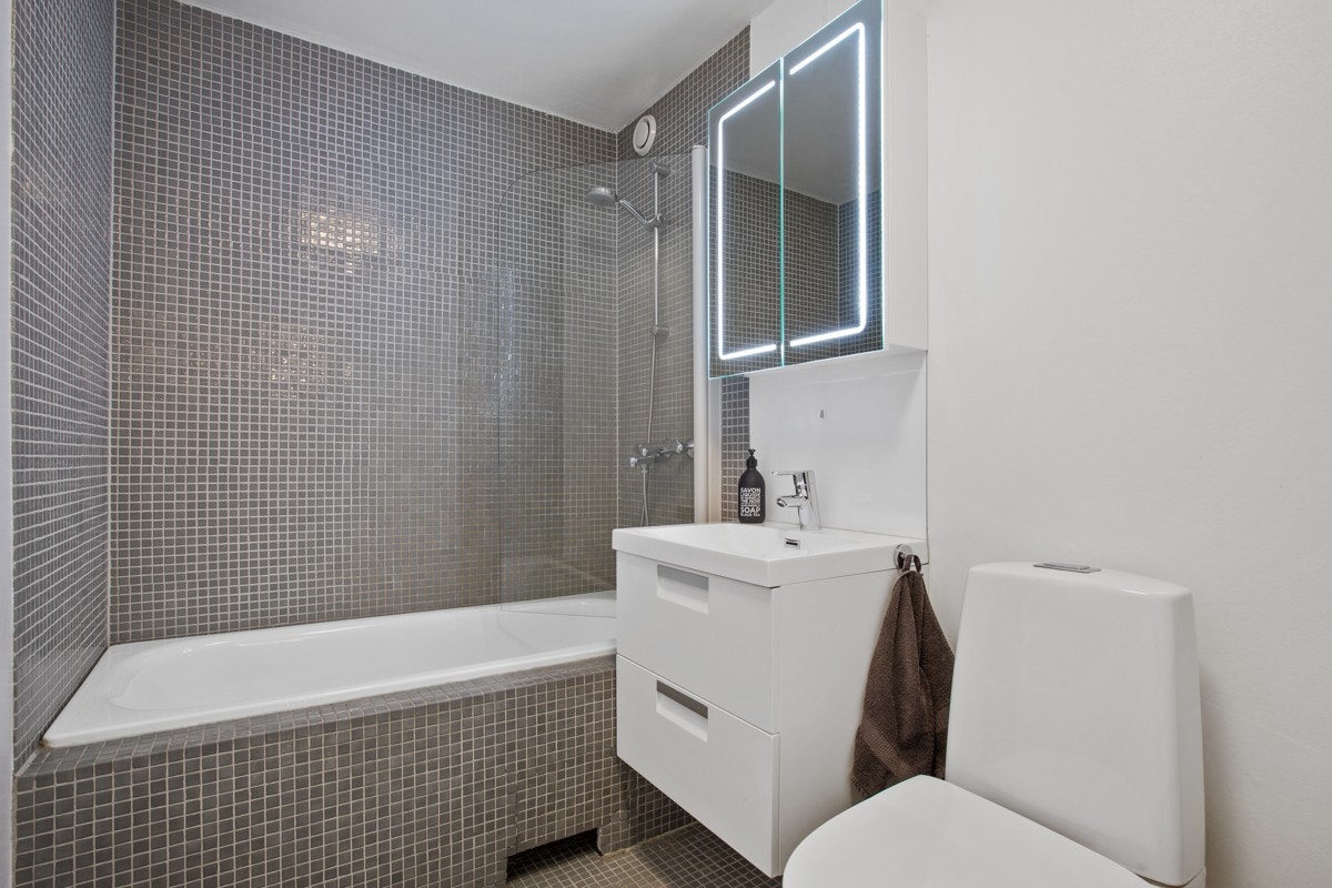 Badet består av innmurt badekar med svingbar dusjvegg, vegghengt servant, vegghengt speilskap og gulvstående toalett. Galleribilde