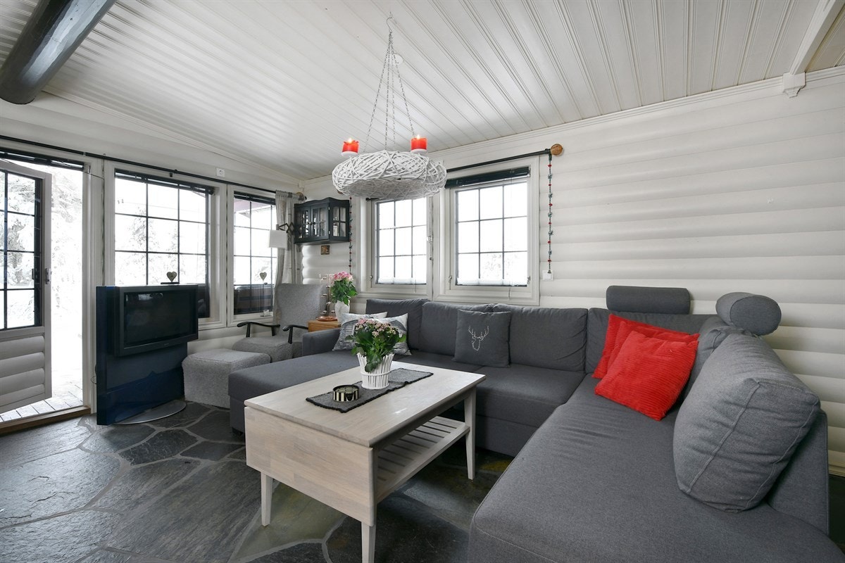 Hytta har en nydelig stue med ekstra takhøyde, store vindusflater, hvitmalt panel og en flott, mørk takbjelke. Galleribilde
