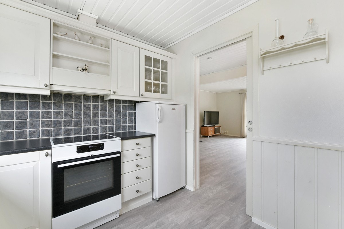 Innredningen hvite profilfronter, vitrineskap, åpen krydderhylle, sort laminatbenkeplater, og nedfelt beslag med vaske- og skyllekum. Videre har kjøkkenet ventilator over kokesonen, integrert oppvaskmaskin, nisje for komfyr og plass for kjøleskap. Galleribilde